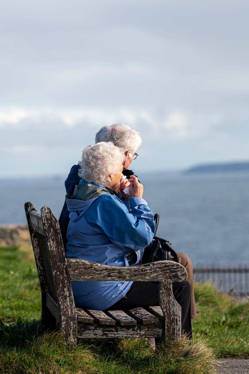Mann und Frau sitzen tagsüber auf einer grauen Holzbank und blicken auf das blaue Meer
