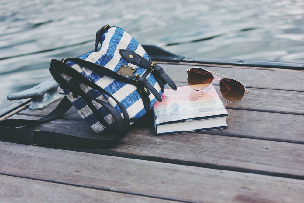 blue and white striped crossbody bag near book and sunglasses on sea dock during daytime