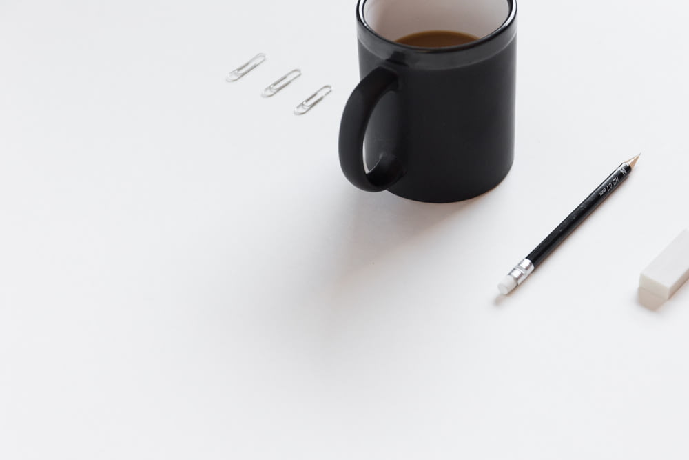 tres clips blancos, una taza de cerámica negra, un lápiz negro y un borrador de lápiz blanco sobre una superficie blanca