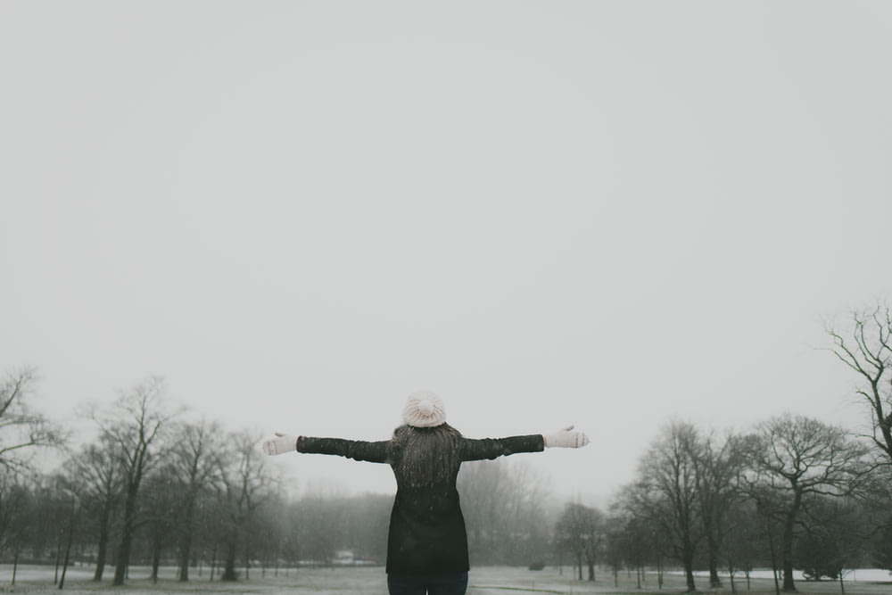 woman in black long-sleeved shirt standing and facing trees with stretched arms under white sky