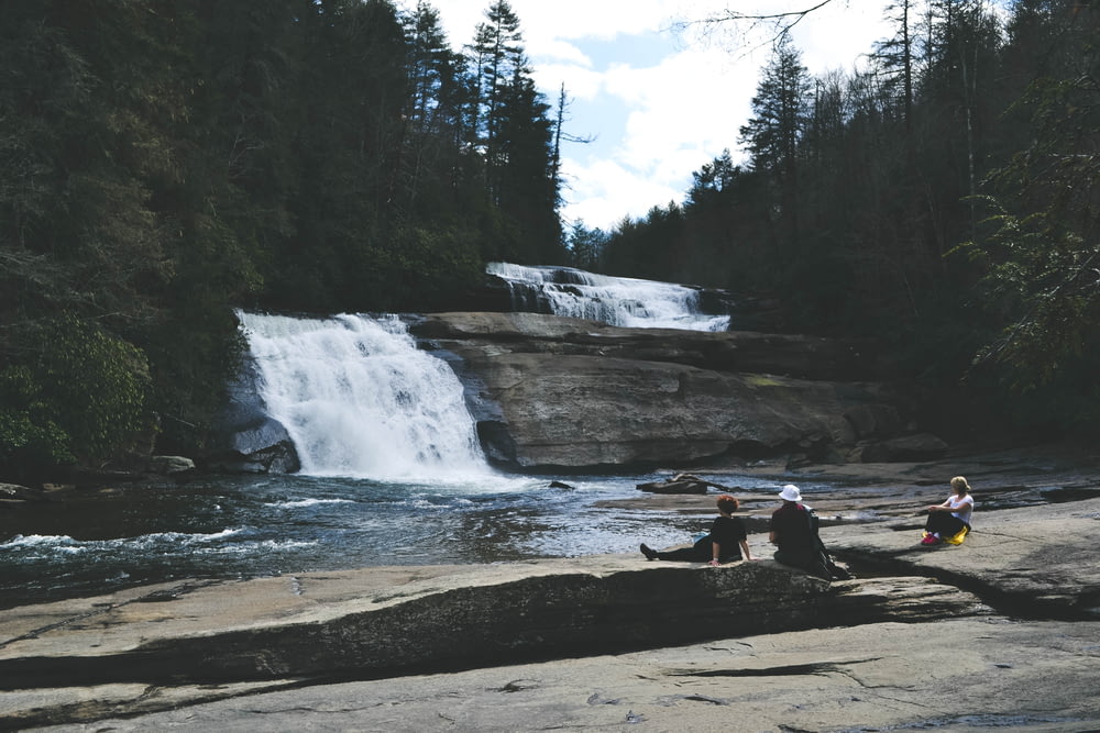 three people sitting on gray surface in front of waterfall