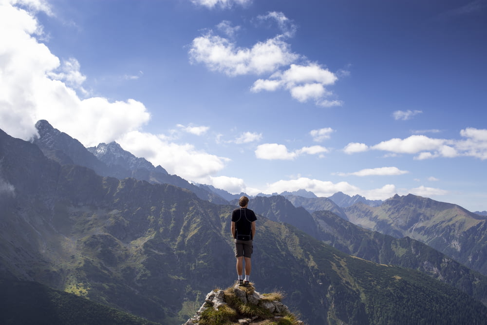 Mann trägt schwarzes Hemd und graue Shorts auf Berghügel neben Bergen unter weiß-blau bewölktem Himmel