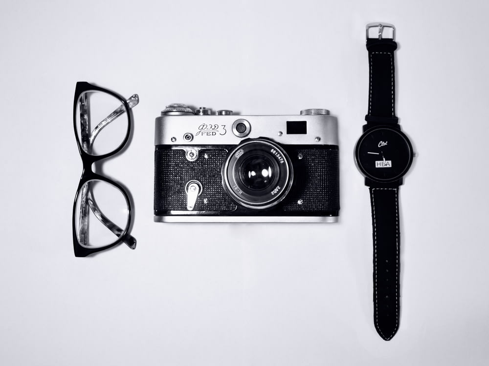black and silver camera; eyeglasses with black frames; black analog watch