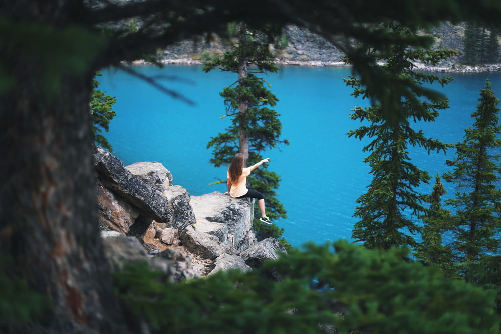 girl sitting on cliff near trees pointing towards body of water