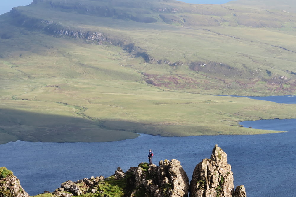 uomo in piedi in cima alla formazione rocciosa accanto al mare vicino alle montagne