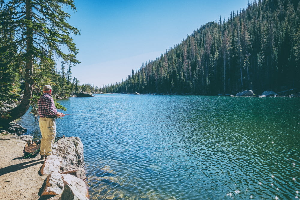 hombre de pie sobre la roca frente al lago rodeado de árboles durante el día