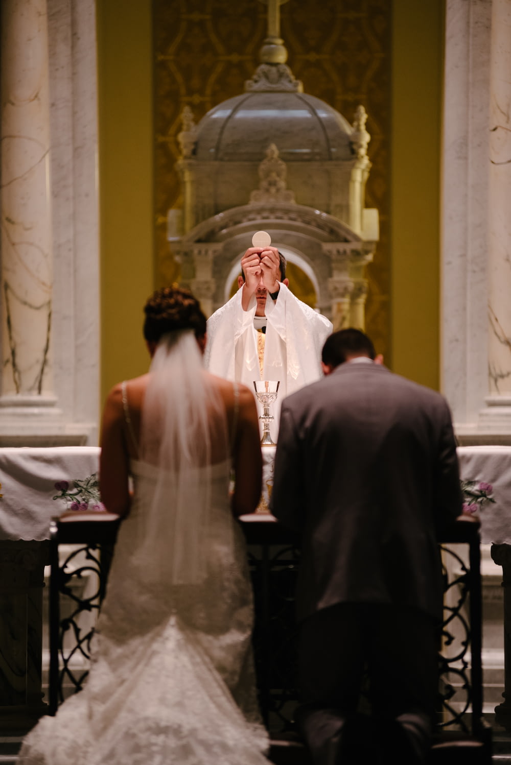 sposo e sposa inginocchiati davanti al sacerdote che alza il Santissimo Sacramento