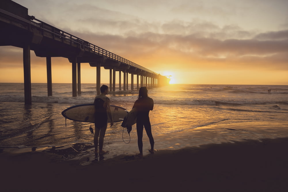 日没時に人の横でサーフボードを運ぶ女性