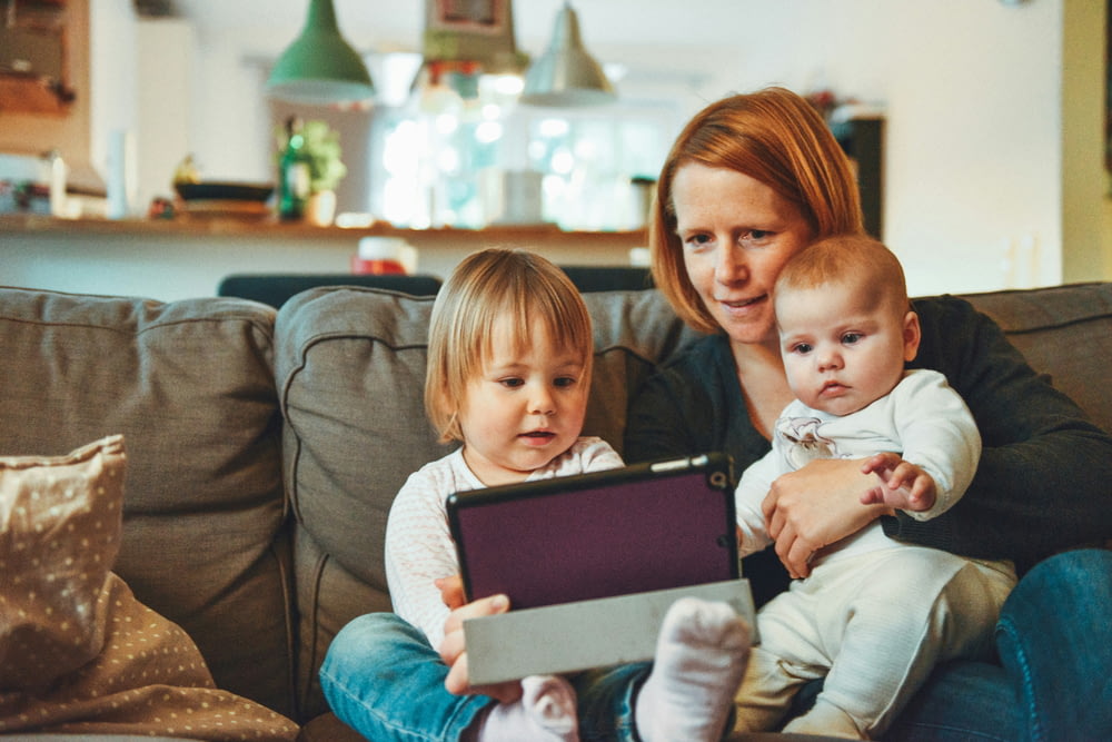 deux bébés et une femme assis sur un canapé tout en tenant un bébé et en regardant sur une tablette