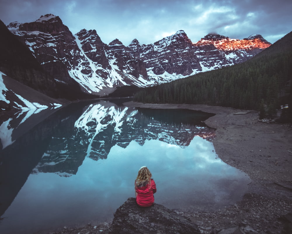 Femme assise sur un rocher en face d’un plan d’eau calme et d’une vue sur les montagnes