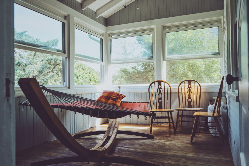 photographie de l’intérieur d’une maison en bois gris, blanc et brun avec trois chaises Windsor en bois brun à côté d’un hamac brun avec des fenêtres en verre