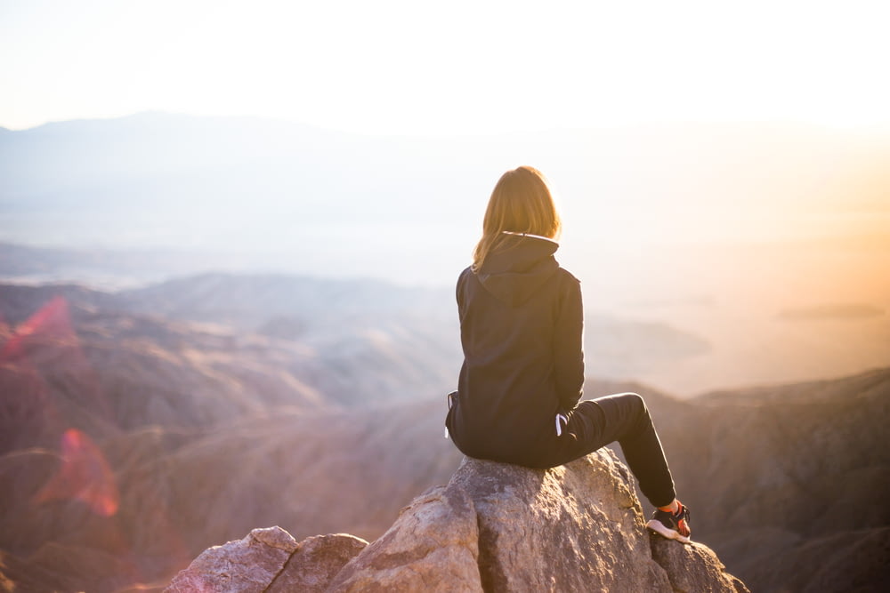 pessoa sentada no topo da rocha cinza com vista para a montanha durante o dia