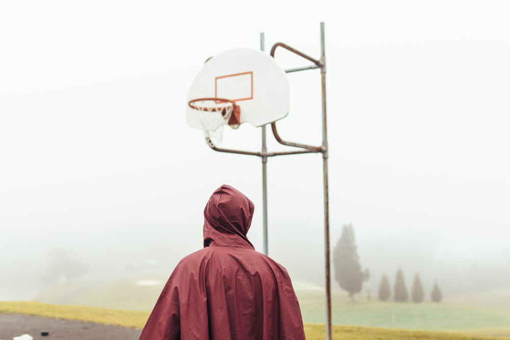 안개 낀 낮에 흰색과 회색 농구 골대 아래에 서 있는 적갈색 비옷을 입은 사람