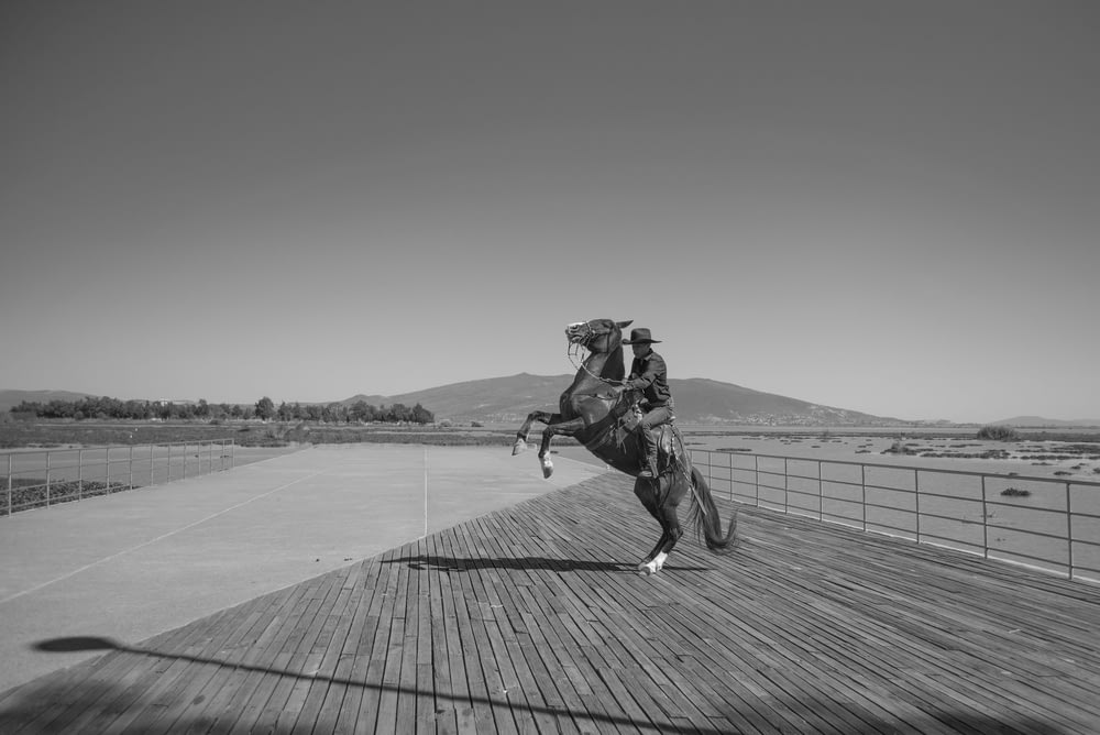 grayscale photo of man in jacket and pants riding horse