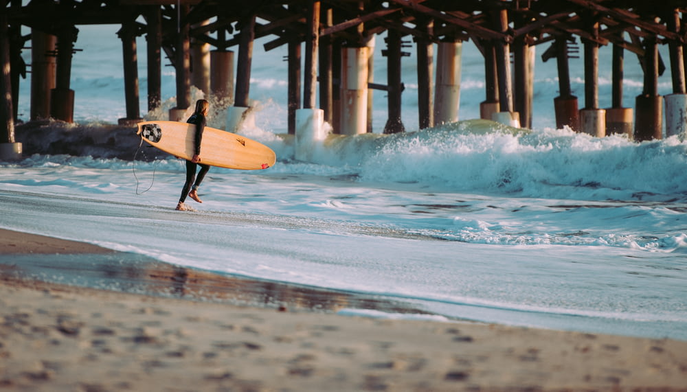 木製の波止場近くの波の手前、海岸に立つサーフボードを持つ人