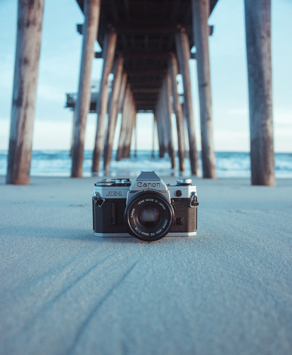 Fotocamera Canon AE-1 nera e grigia sulla sabbia grigia sotto il molo marrone vicino allo specchio d'acqua durante il giorno