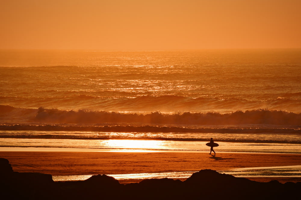person holding surfboard walking on seashore during golden hour