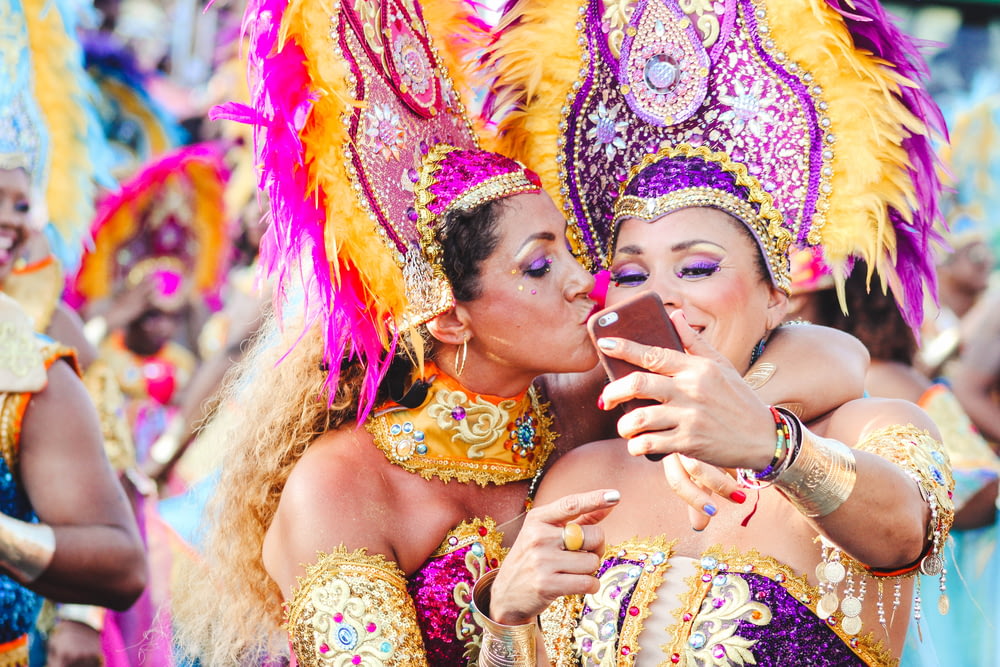 2人の女性カーニバルダンサーが自撮りのポーズをとり、1人がもう1人の頬にキスをします。