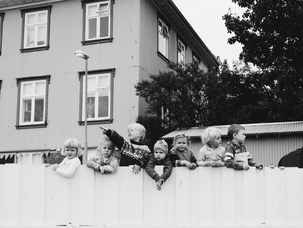 grayscale photo of seven children standing in wooden fence near house