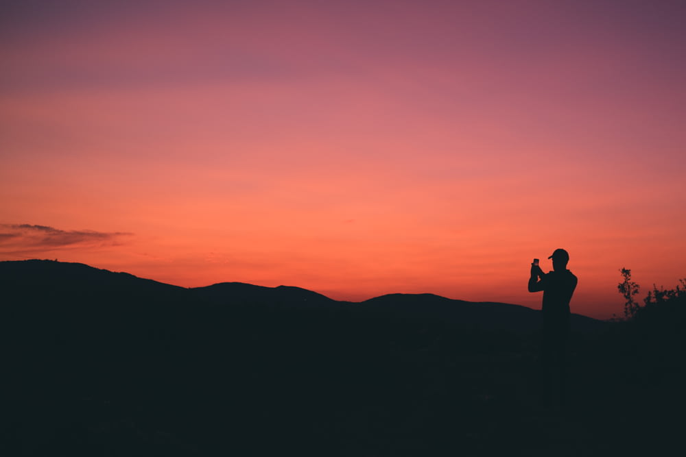 silhouette of person taking photo of mountain during sunset
