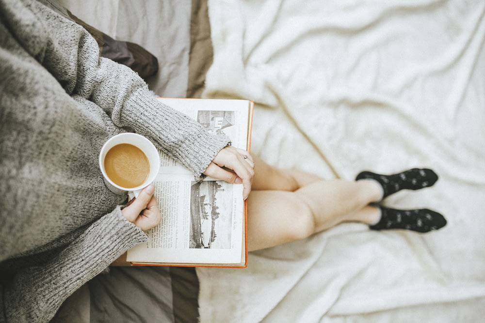 Femme tenant une tasse de café dans la main droite et lisant un livre sur ses genoux tout en le tenant ouvert avec sa main gauche dans une pièce bien éclairée