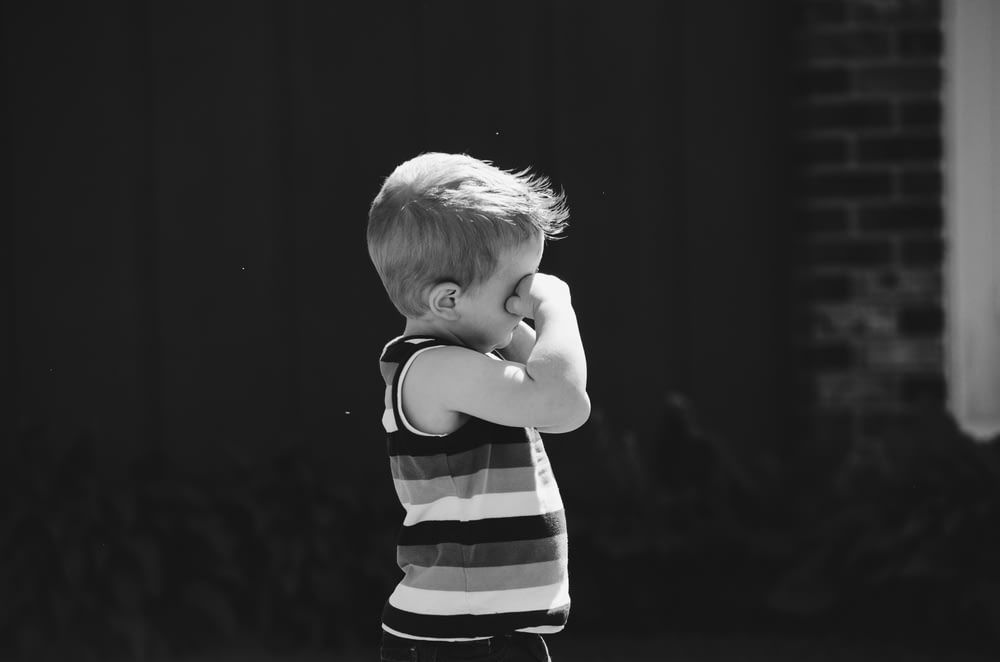 Graustufenfoto eines Jungen, der ein Auge bedeckt, während er hinter einer Betonwand steht