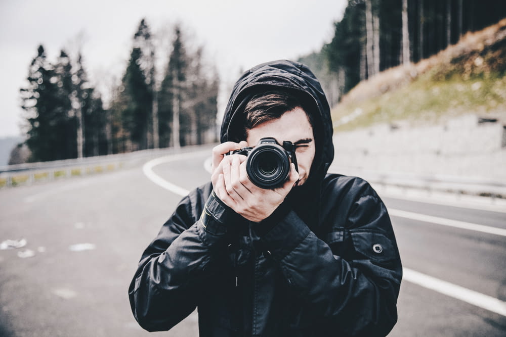 man in black jacket taking picture with DSLR camera on road