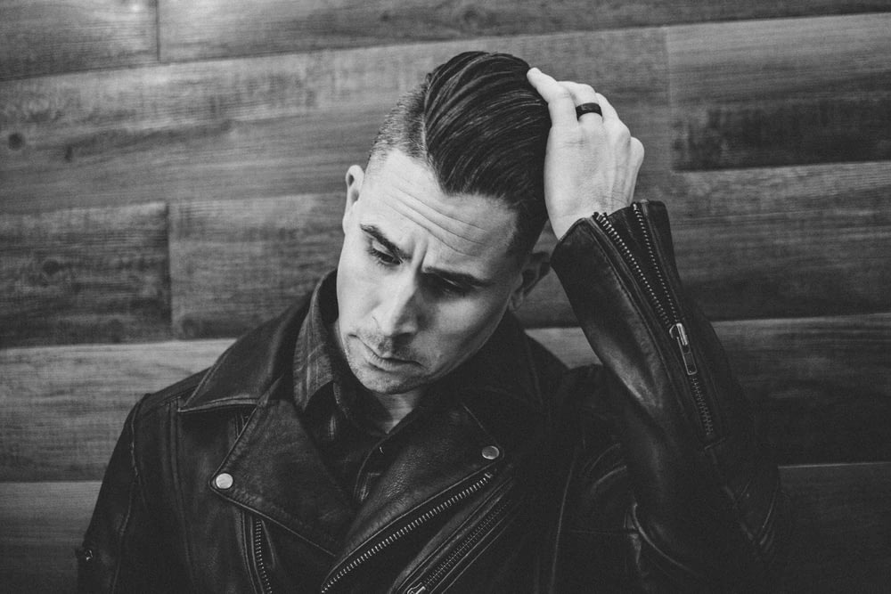 grayscale photography of man wearing leather jacket leaning against wall while fixing hair