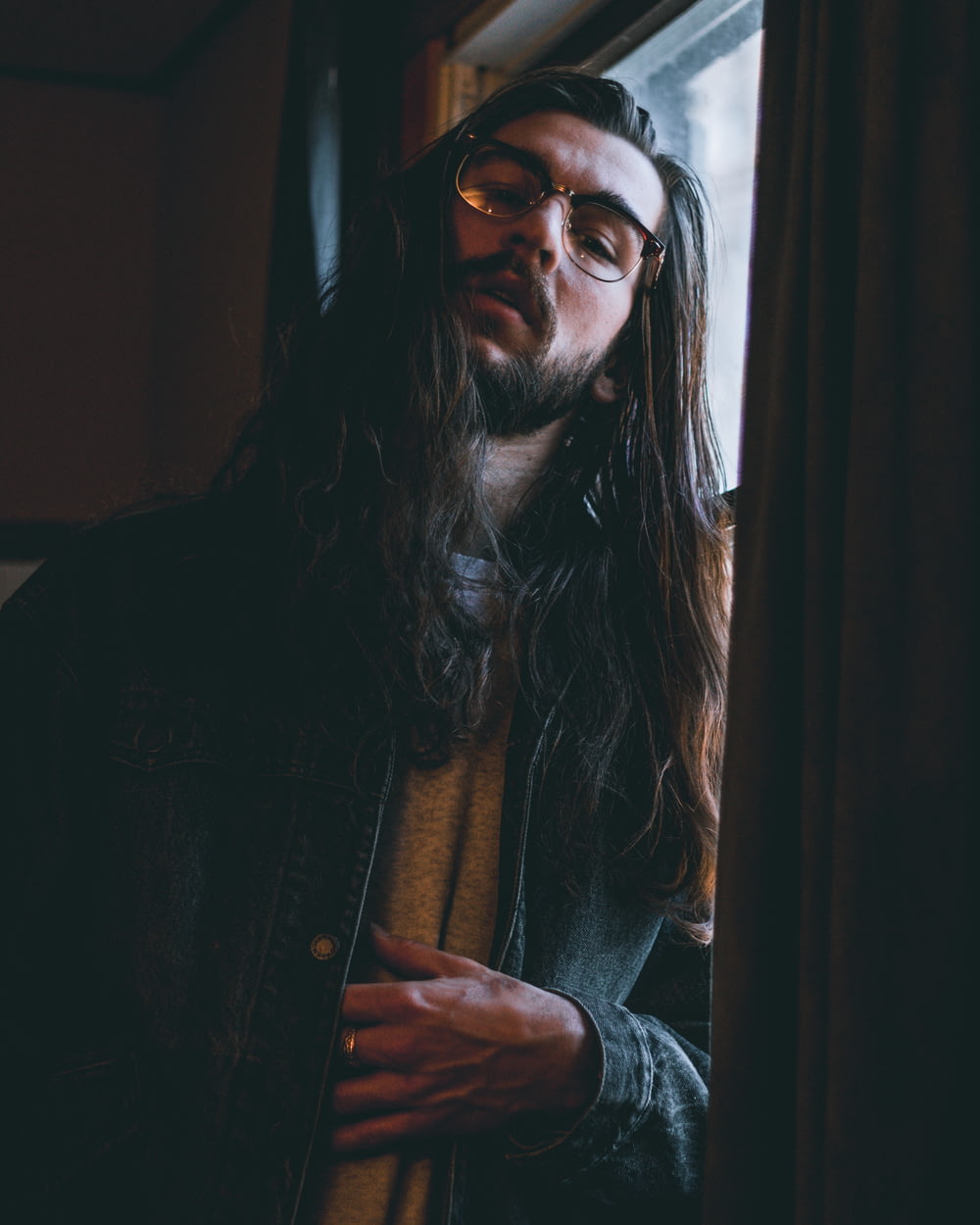 long haired man wearing eyeglasses and blue denim jacket leaning on window