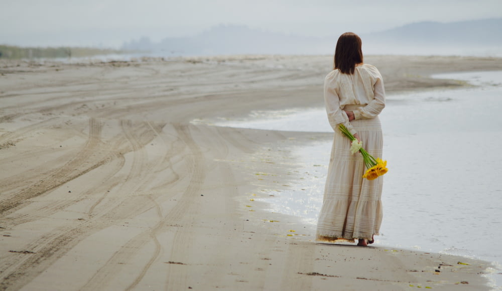 woman holding yellow petaled flowers while standing near seashore