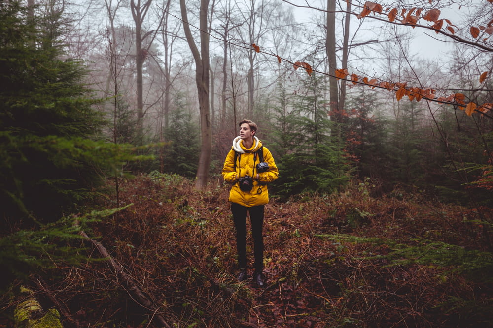 man in yellow jacket holding black camera standing on brown grasses under trees