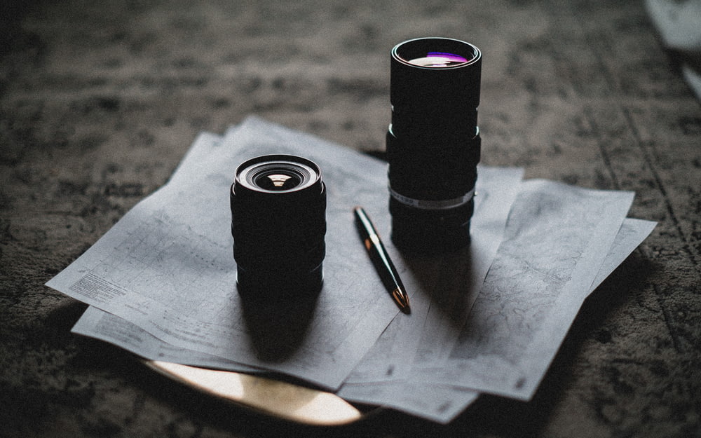 two camera lens on top of white printed papers and black and gold click pen