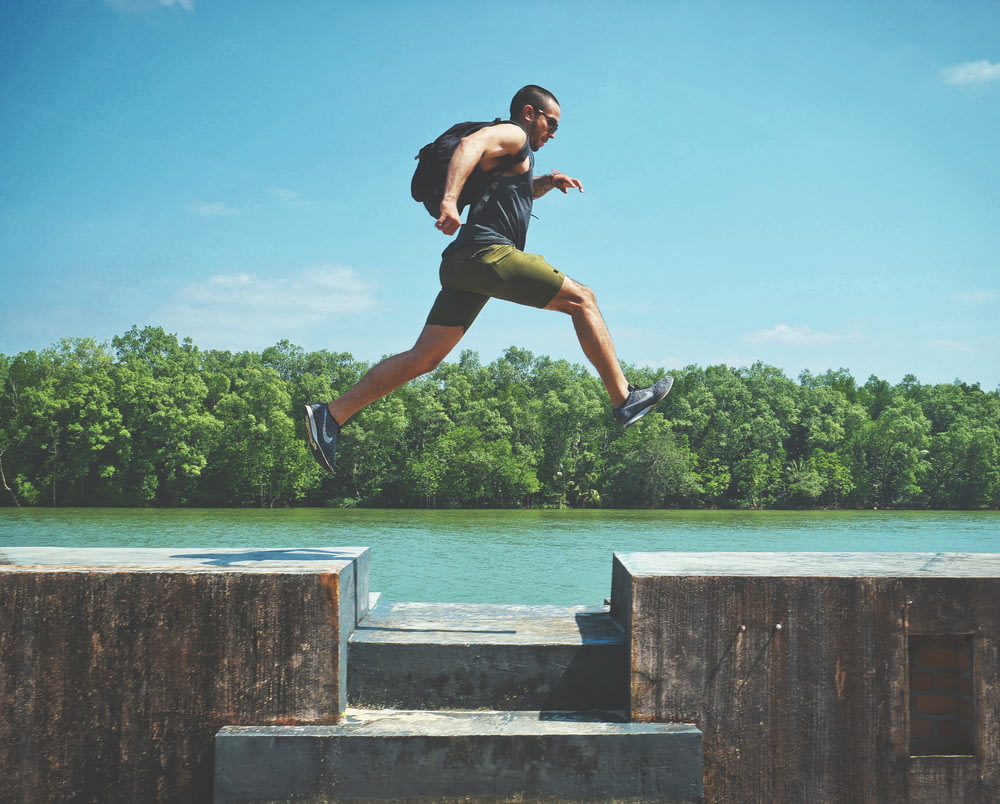 homem saltando na superfície de concreto perto do corpo de água e da floresta à distância durante o dia