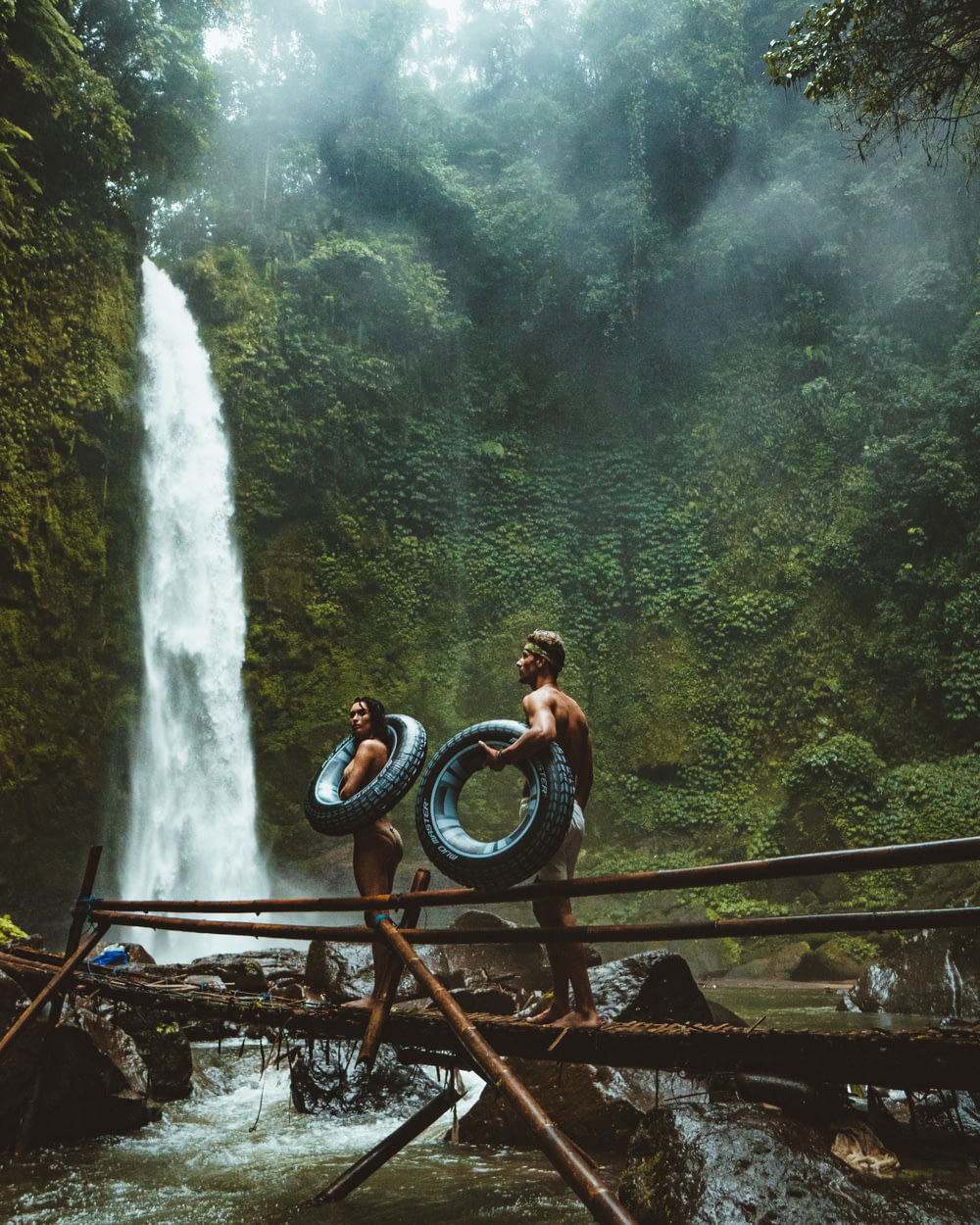 photo of man and woman holding tires and walking on bridge near waterfalls