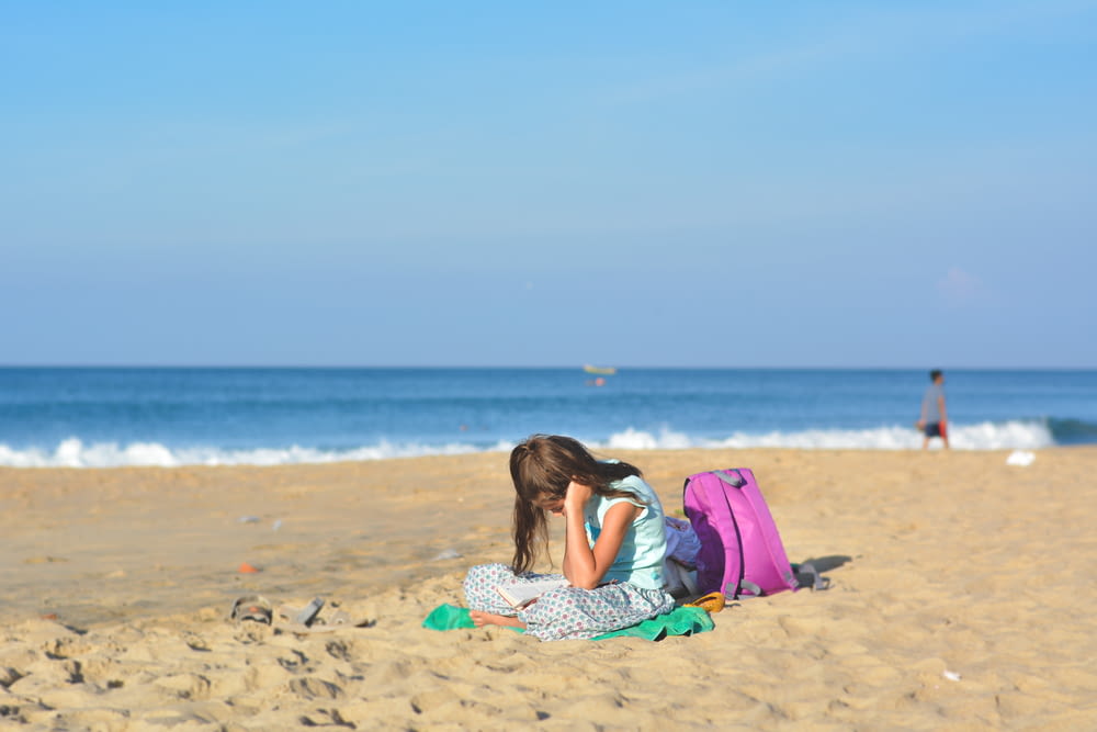 girl sits on shore near purple backpack under blue sky