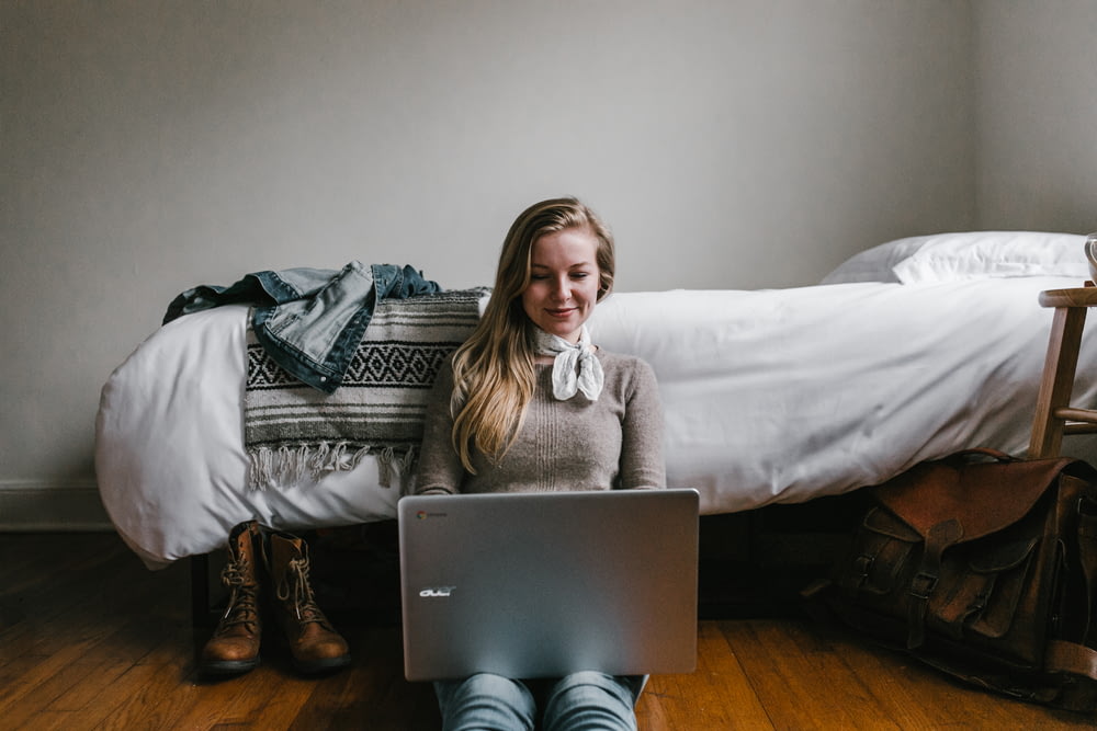 Mujer sentada al lado de una cama mientras usa una computadora portátil
