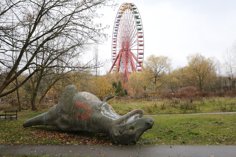 statua di dinosauro in cemento grigio sdraiata sull'erba verde vicino alla ruota panoramica rossa