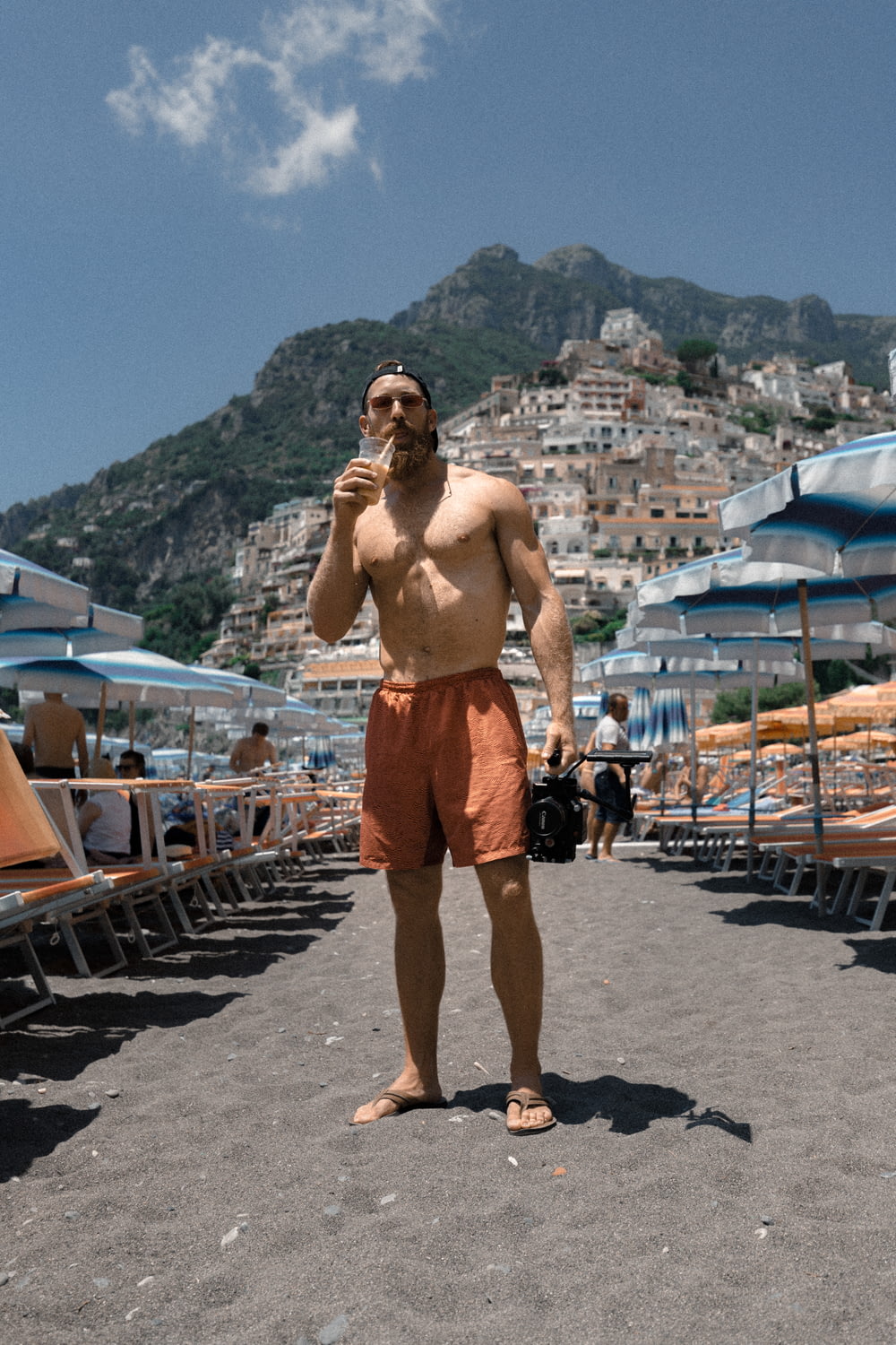 オレンジ色のショートパンツを履いた男がビーチの砂の上に立ち、カップで飲む