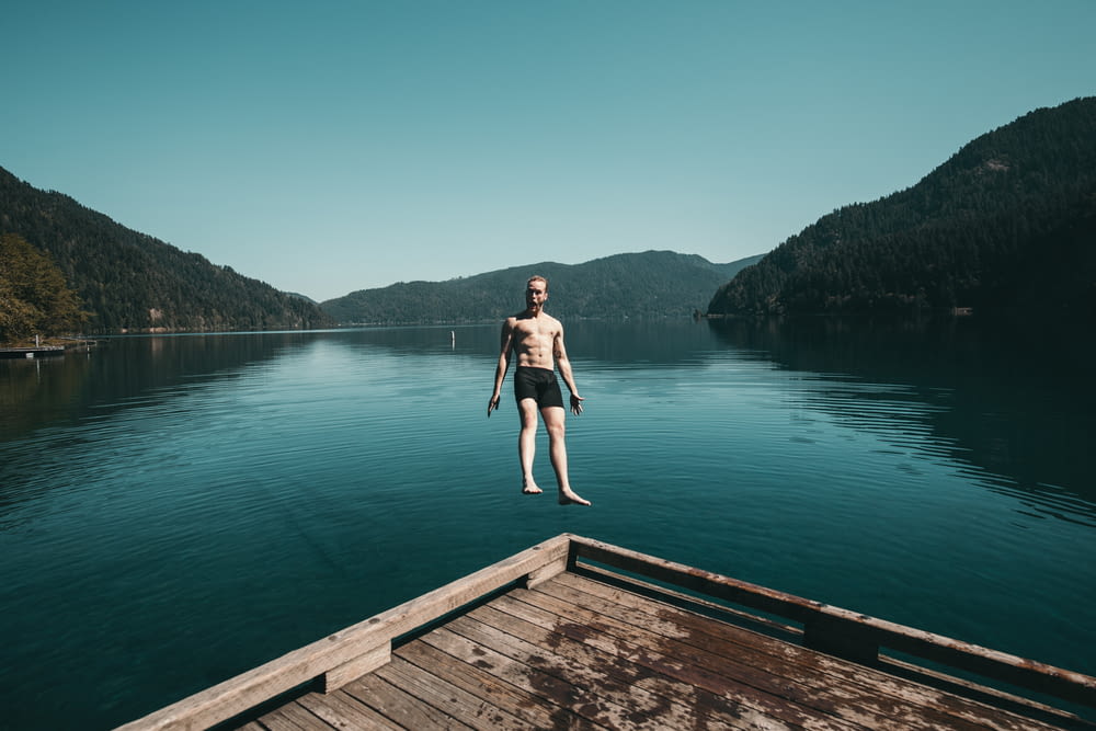 man jumping from brown wooden platform unto lake facing backwards