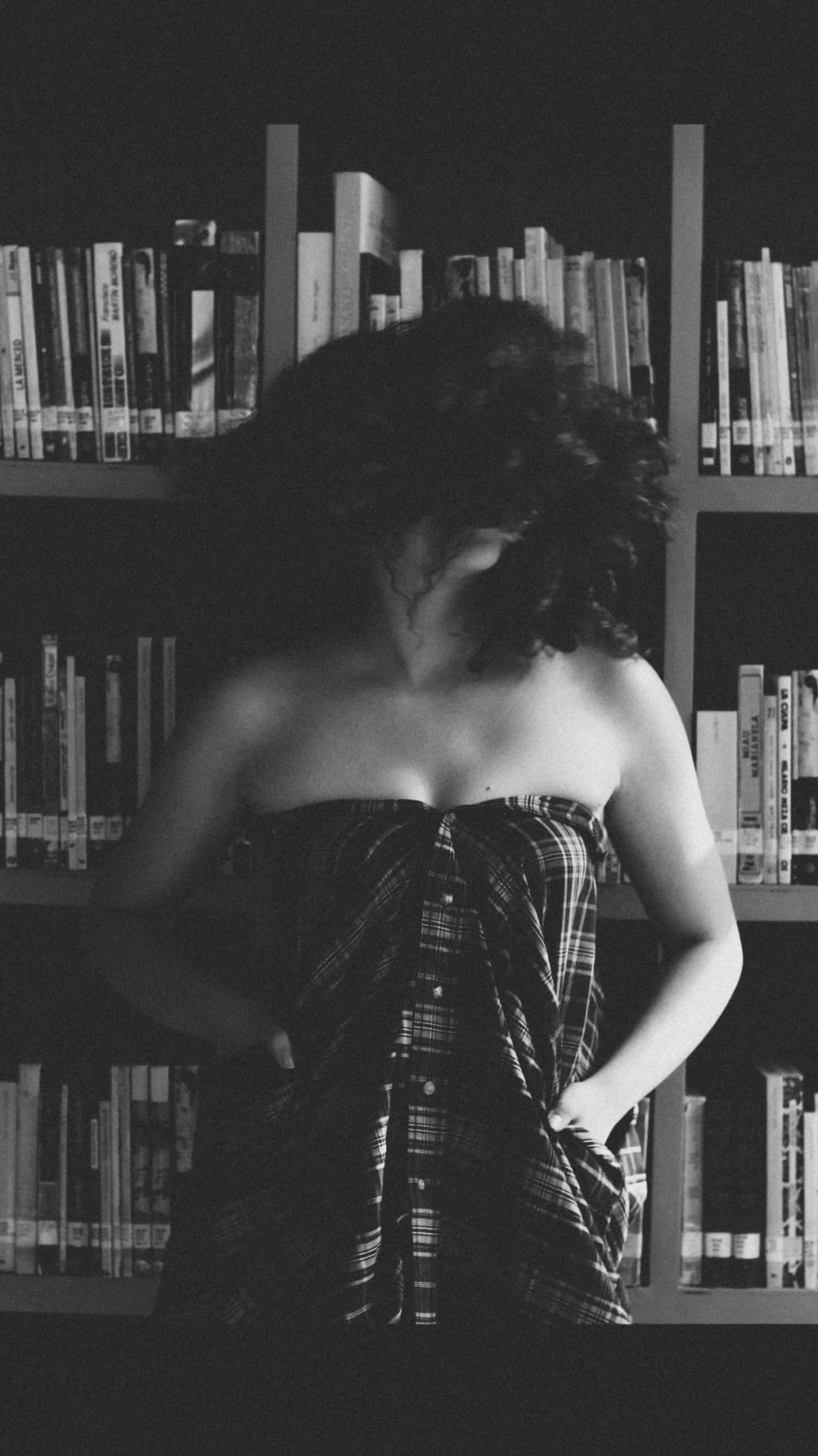 grayscale photography of woman wearing brown bottoms with curly hair standing beside bookshelf