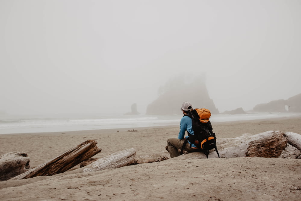 man wearing orange and black hiking backpack sitting on log on shore looking at distance during daytime