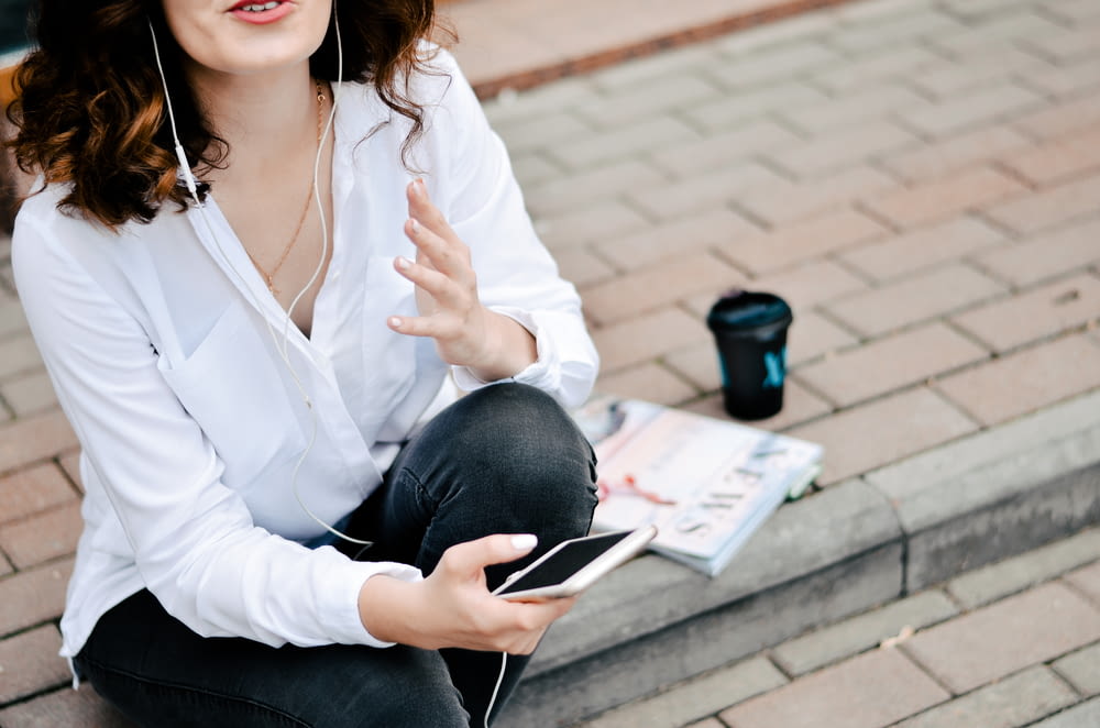 woman holding smartphone while sitting on brick pavement during daytime