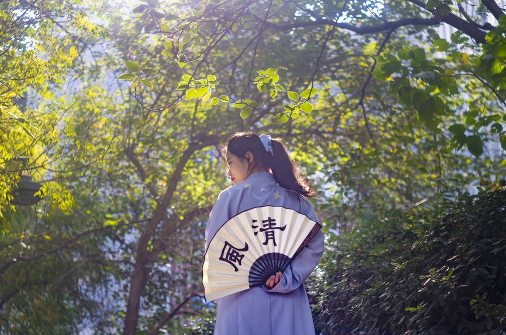 woman holding white and black hand fan standing beside green trees during daytime