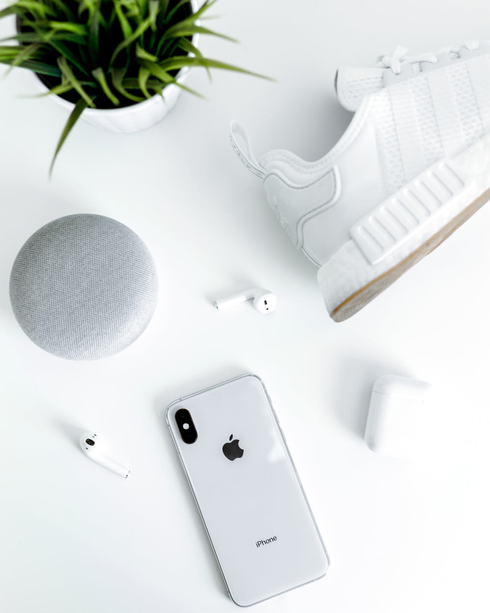 silver iPhone X near white adidas NMD shoe, AirPods with case, and chalk Google Home Mini
