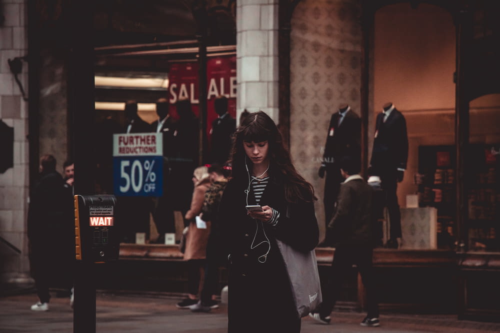 standing woman wearing black coat using smartphone and earphones