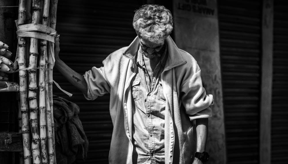 grayscale photography of standing man wearing jacket holding tied bamboos