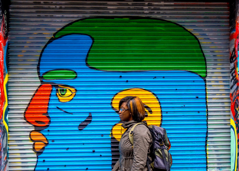 회색 재킷을 입은 여자가 파란색, 주황색, 녹색 얼굴 벽화 근처를 걷고 있습니다