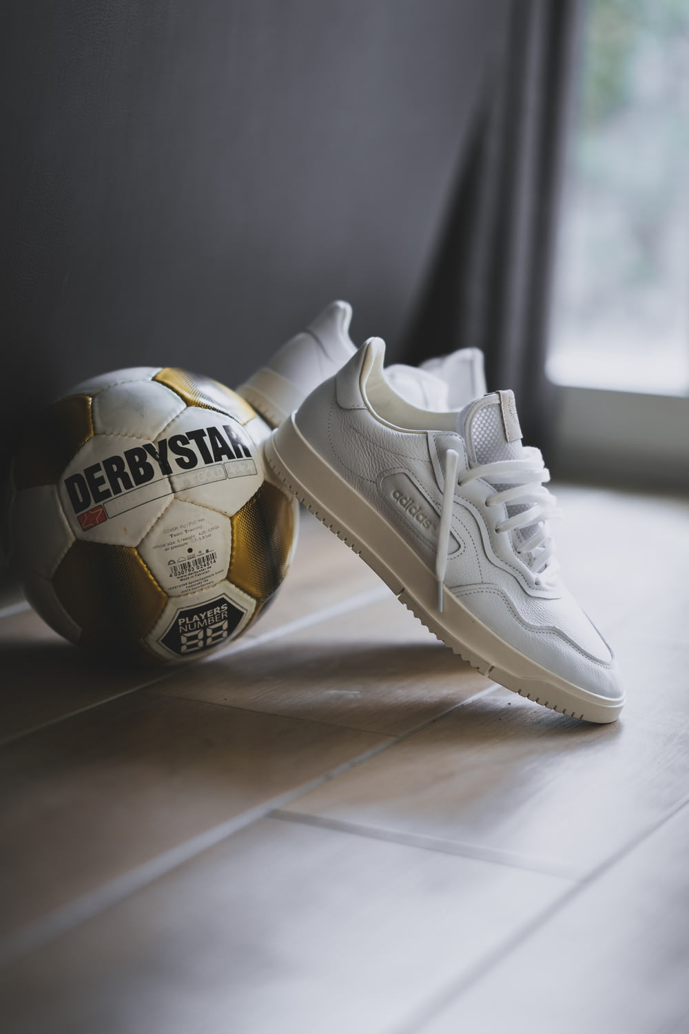 pair of white Adidas low-top sneakers beside Derbystar soccer ball