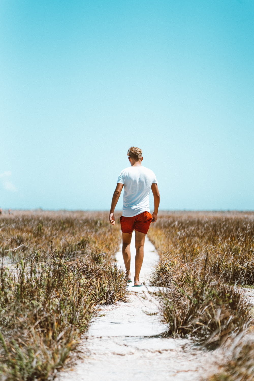 uomo in camicia bianca e pantaloncini rossi che cammina sulla sabbia bianca tra le erbe