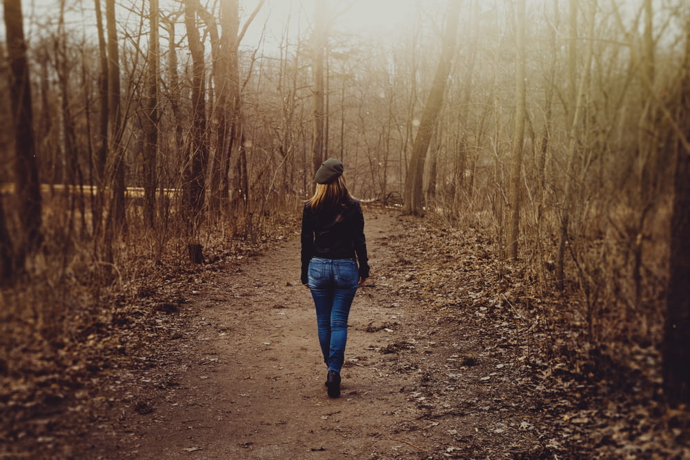 woman in black sweater and blue denim skinny jeans walks on dirt road beside leafless trees